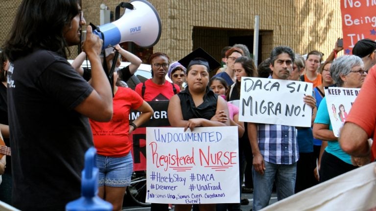 A woman speaks through a bullhorn, immigrants hold signs, listen to her speak during a rally