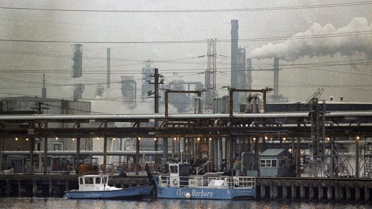 two clean harbor cleanup boats work in a waterway alongside an industrial refinery, smoke stacks in the background