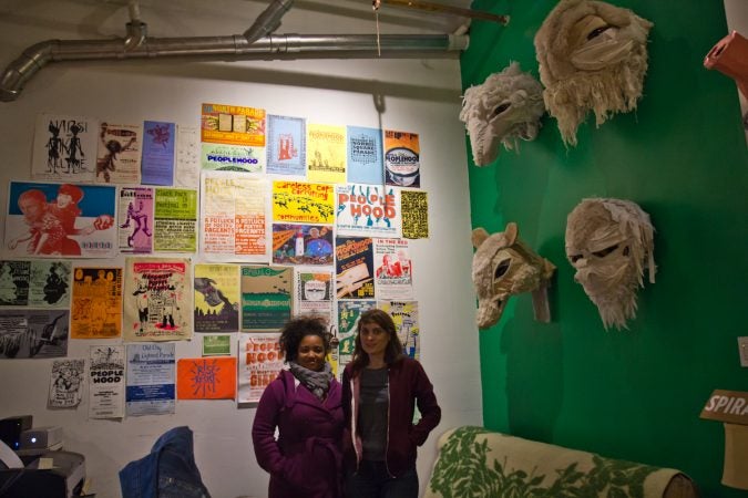 Spiral Q Artistic Directors Jennifer Turnbull and Liza Goodell stand before posters of Peoplehood Parades past, and giant puppet heads at their warehouse space in West Philadelphia. (Kimberly Paynter/WHYY)