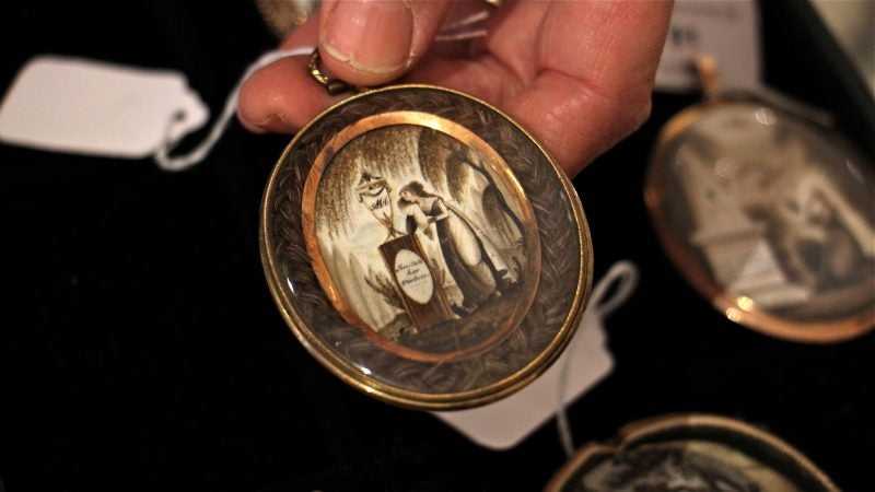 A palm-sized oval pendant holds a painting on ivory of a woman grieving beside a gravestone. The image is framed by braided human hair.