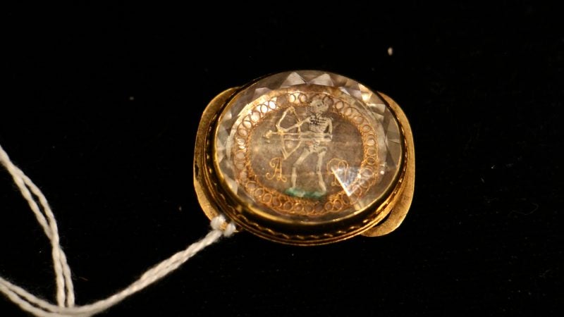 A mourning slide, intended to be worn on a ribbon, depicts a skeleton holding a bow and arrow and the initials of the deceased visible through a faceted rock crystal.