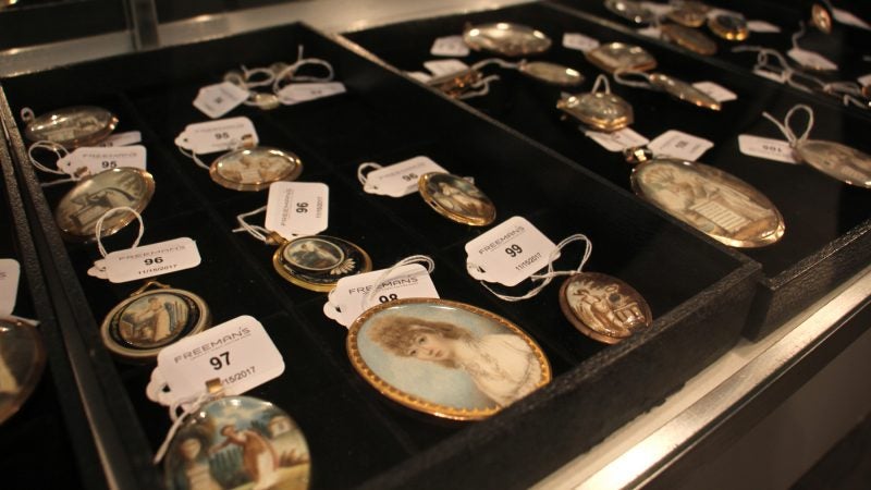 Lockets depicting various emblems of mourning are displayed in a glass case with auction tags attached