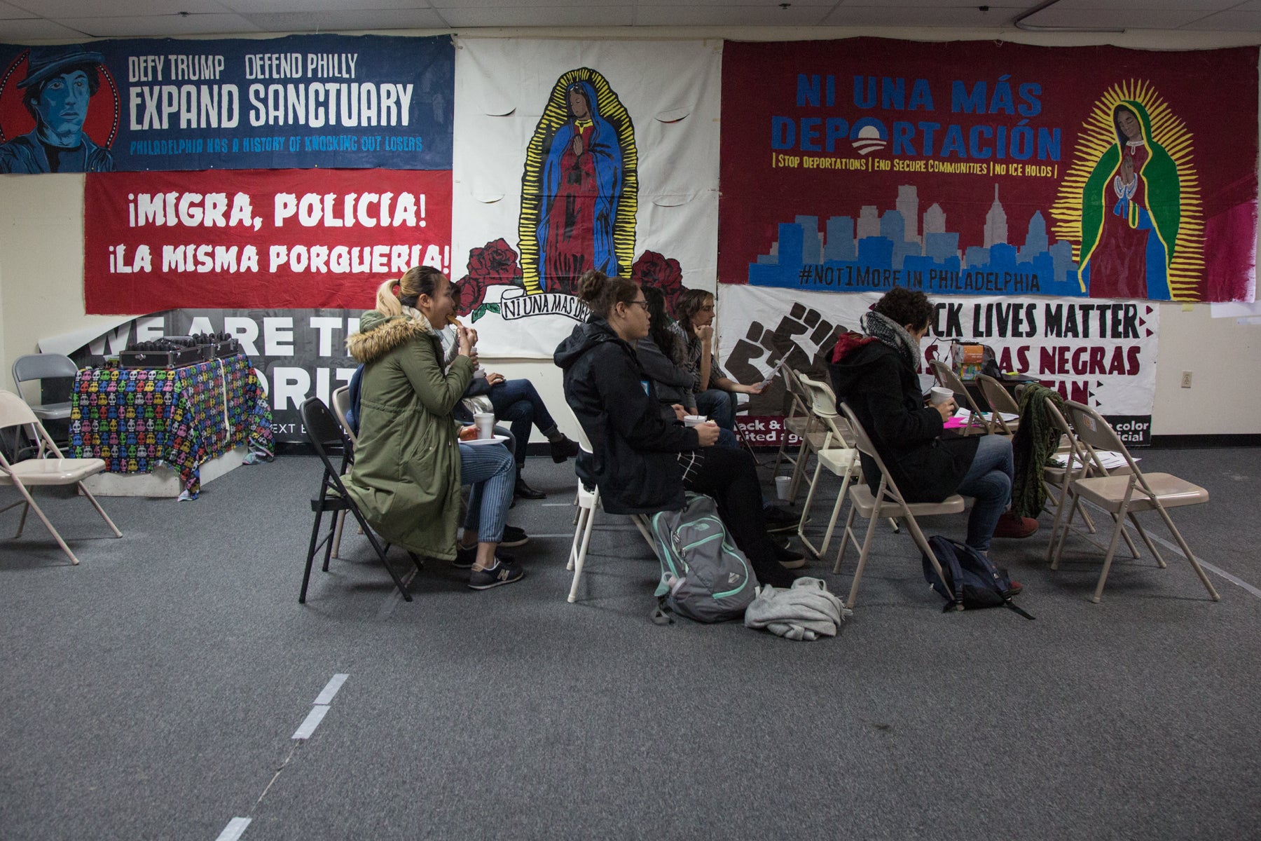 People in winter coats sit on folding chairs against a background of colorful posters in Spanish language