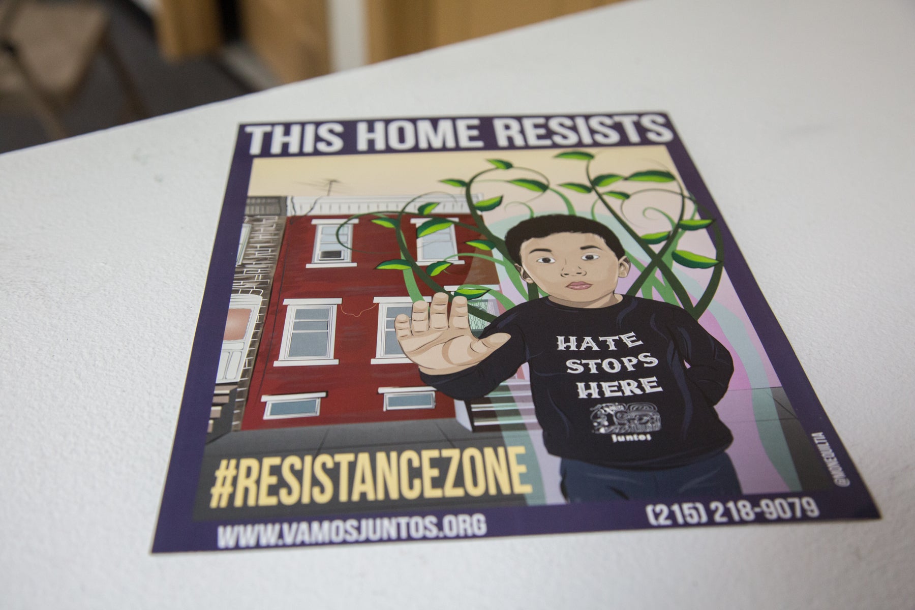 A poster shows a boy standing in front of a rowhome with his hand held up palm out. Across the top it says "this home resists."