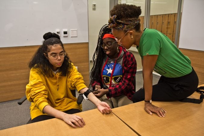 Medical student Camille McCallister supervises Syre students Nydia Henry (right) putting a tourniquet on Danny Miles (left). (Kimberly Paynter/WHYY)