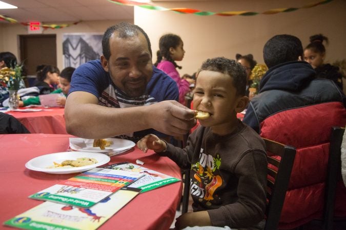 Nelson D'Jesus playfully feeds his son, Mikeli,4, a sweet at APM's Three Kings Day/Octavious celebration for diplaced families from Puerto Rico in North Philadelphia on January 12 2018. (Emily Cohen for WHYY)