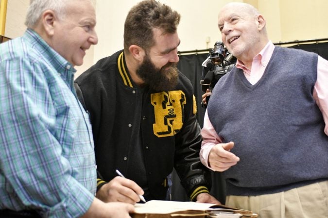 Alumni and volunteers Chuck Steinburg (left) and David Kahn (right) share the history of the guestbook as Philadelphia Eagles center Jason Kelce leaves his signature. The entries in Central High’s guestbook date back to 1838 and include two presidents as well as other notable alumni. (Bastiaan Slabbers/for WHYY)