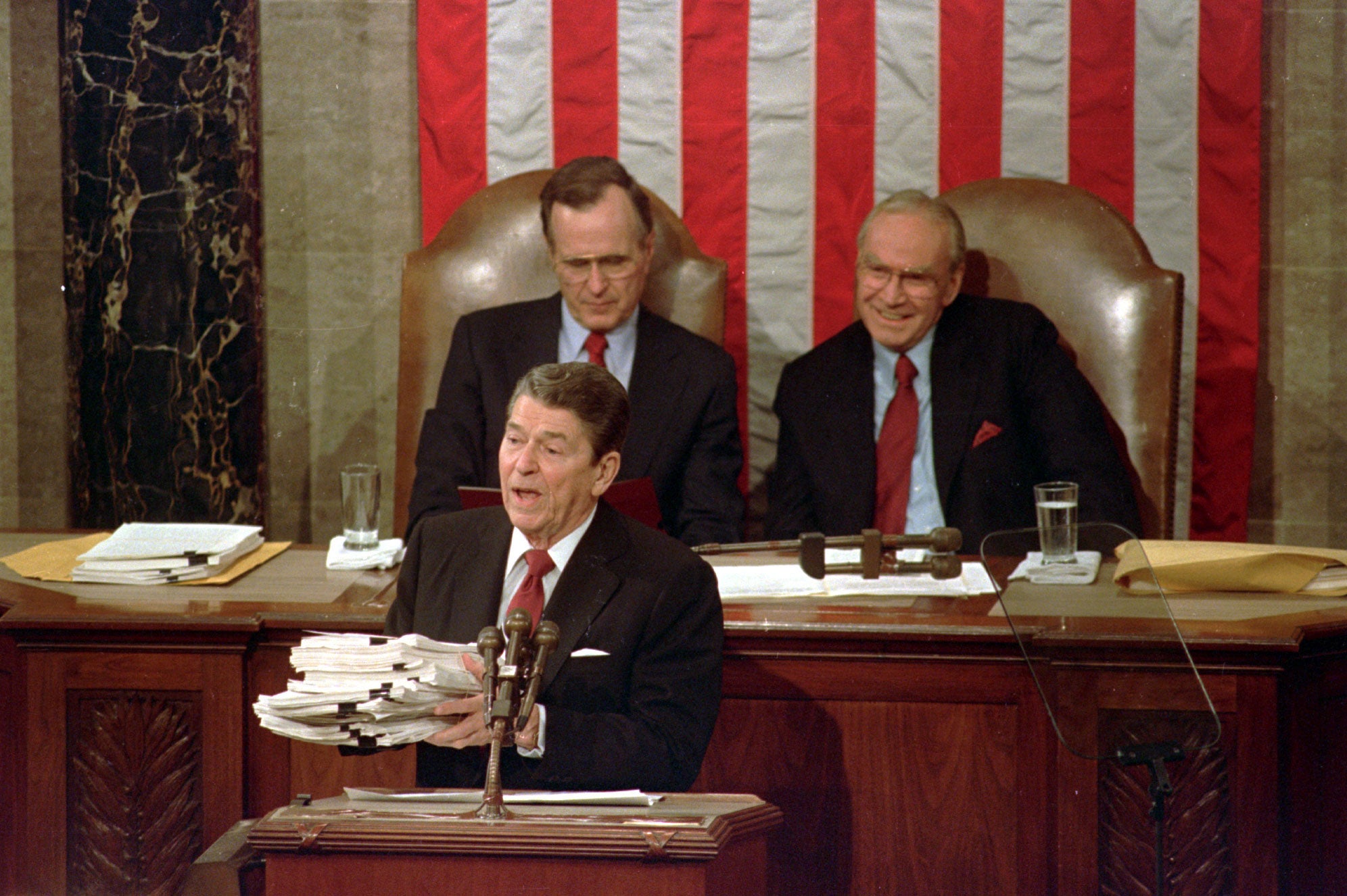 During his 1988 State of the Union address, President Ronald Reagan holds up a 14-pound continuing resolution for the budget, part of a total package weighing 43-pounds, which the president said was two months late from Congress. Vice President George H.W. Bush (left) and House Speaker James Wright of Texas listen behind him