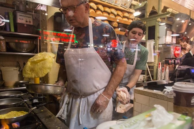 Head chef, Andy Wash, prepares some omelets as Ian McClendon, 23, sneaks in behind to refresh his cheese supply. (Emily Cohen for WHYY)