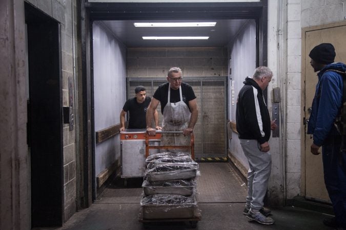 Andy Wash, center, brings up  six double pans and four single pans of Pastrami which has been cured in-house in the market basement, as Nate Ventura, left, follows behind with an extra oven which will be placed inside the deli to help accommodate the demand in preparation for the anticipated influx of customers coming from the first day of the Philadelphia Flower Show the next day. (Emily Cohen for WHYY)