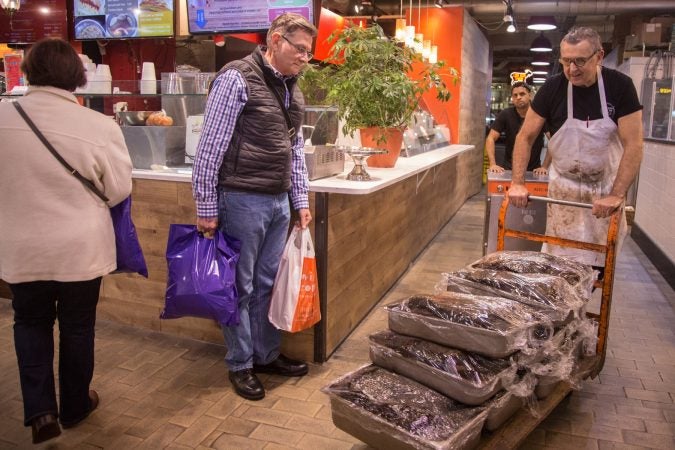 A market patron watches as Andy Wash, right, brings up six double pans and four single pans of Pastrami which has been cured in-house in the market basement, as Nate Ventura, behind, follows behind with an extra oven which will be placed inside the deli to help accommodate the demand in preparation for the anticipated influx of customers coming from the first day of the Philadelphia Flower Show the next day. (Emily Cohen for WHYY)