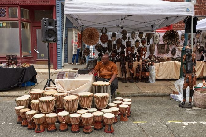 DJ Master JOE Love sits beside African Djembes and figurines for sale at Odunde Festival in Philadelphia on Sunday, June 10, 2018. These goods are made in Senegal, West Africa. (Natalie Piserchio for WHYY News) 