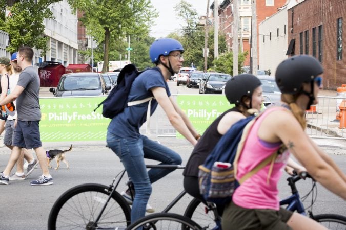 People on bikes and on foot pass in front of barriers that are blocking Broad Street from traffic as a part of the third annual Philly Free Streets event on August 11, 2018. (Rachel Wisniewski for WHYY)