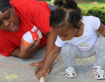 Marletha Muhammad helps her daughter, Khanila, spell her name with chalk at a June playgroup meeting in Rocky Mount, North Carolina The purpose of the meetings is to encourage healthy interactions between parents and their young children. (Liz Bell/The Hechinger Report)
