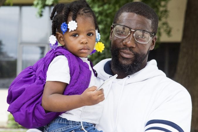 Warren Kale, 42, holds his daughter Katie, 2, outside of the Made in America festival on the Benjamin Franklin Parkway Saturday. Kale and his family have been homeless since December and were hoping to sell water bottles outside of the festival in order to help save up for an apartment. Although Kale had previously obtained a permit to vend, he was told Saturday morning 1 that his permit was invalid. (Rachel Wisniewski for WHYY)