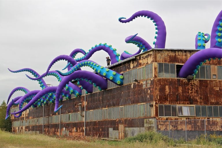 Giant purple green and blue tentacles protrude from the windows of a dilapidated warehouse at the navy yard