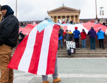 A man wears a cape made from a Puerto Rican during an event to remember the people who died in Hurricane Maria in Puerto Rico. The group of activists gathered on the steps of the Philadelphia Museum of Art on Oct. 13, 2018. (Miguel Martinez for WHYY)