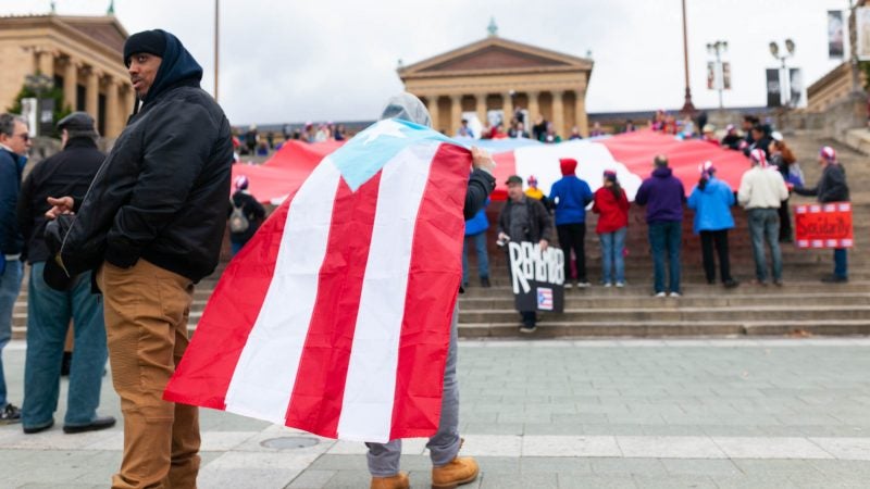 A man wears a cape made from a Puerto Rican during an event to remember the people who died in Hurricane Maria in Puerto Rico. The group of activists gathered on the steps of the Philadelphia Museum of Art on Oct. 13, 2018. (Miguel Martinez for WHYY)