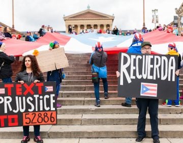 Yecenia Vazquez (left) and Jaime Knowles hold signs in front of a large Puerto Rican flag that was laid it at the steps of the Philadelphia Museum of Art. A group of activist smarched the Benjamin Franklin Parkway to commemorate the people who died in Hurricane Maria in Puerto Rico. (Miguel Martinez for WHYY)