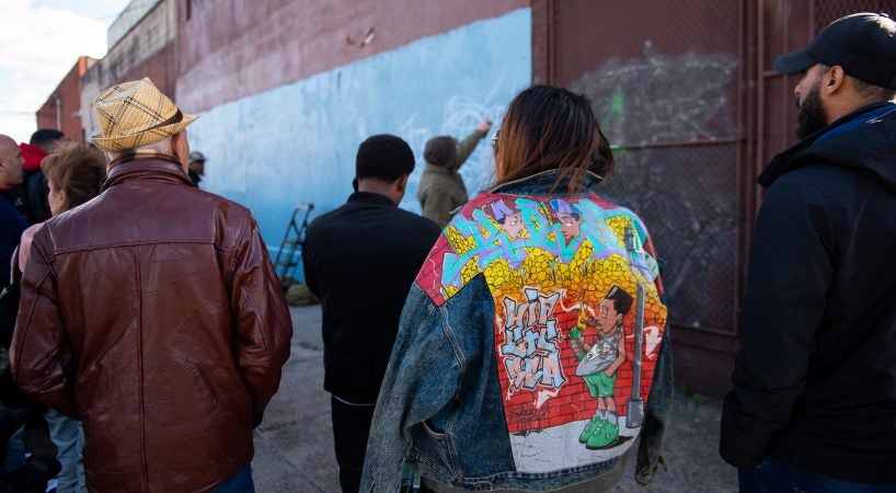 Ashley Sanchez, niece to the late graffiti writer Karaz, wears his jacket while a mural is painted in his honor in Hunting Park on Saturday, November 3, 2018. During large gatherings, like this send off, writers often bring books that they have others sign. (Kriston Jae Bethel for WHYY)