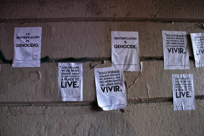 Protest flyers in English and Spanish are pasted on the wall of the Emerald Street railroad overpass, the site of an encampment long used by heroin addicts. (Emma Lee/WHYY)