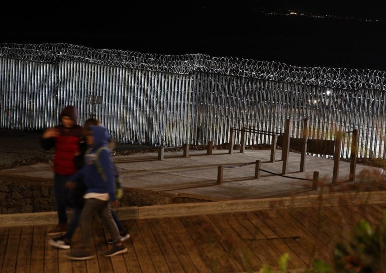 Floodlights from the U.S, illuminate the border wall, topped with razor wire, as a people pass Monday, Jan. 7, 2019, at the beach in Tijuana, Mexico. With no breakthrough in sight, President Donald Trump will argue his case to the nation Tuesday night that a 