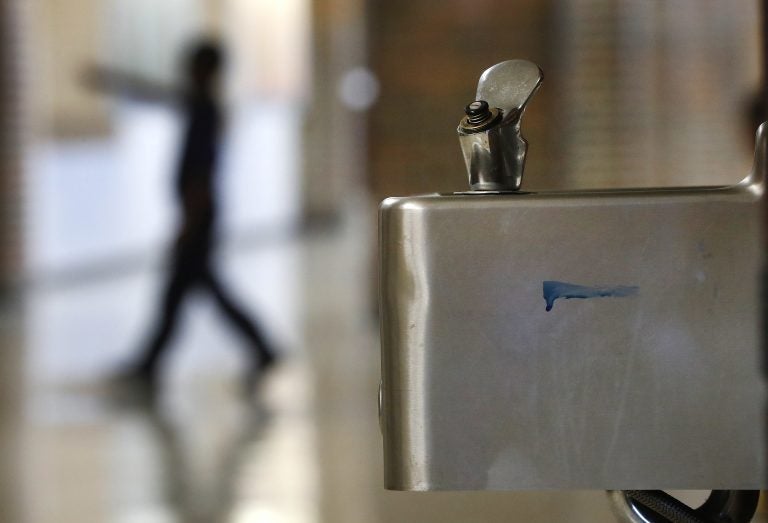 A blurry silhouette of a student is visible in the background, with a up-close sharp image of a water fountain in the foreground.