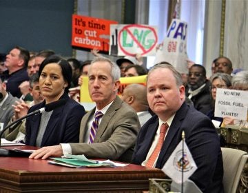 Representatives of Philadelphia Gas Works (from left) Melanie McCottry and President and CEO Craig White, and Matthew Tayler of Liberty Energy Trust, testify before the Committee on Transportation and Public Utilities while opponents raise signs in protest. (Emma Lee/WHYY)