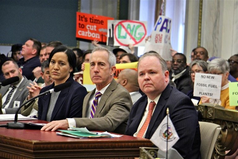 Representatives of Philadelphia Gas Works (from left) Melanie McCottry and President and CEO Craig White, and Matthew Tayler of Liberty Energy Trust, testify before the Committee on Transportation and Public Utilities while opponents raise signs in protest. (Emma Lee/WHYY)