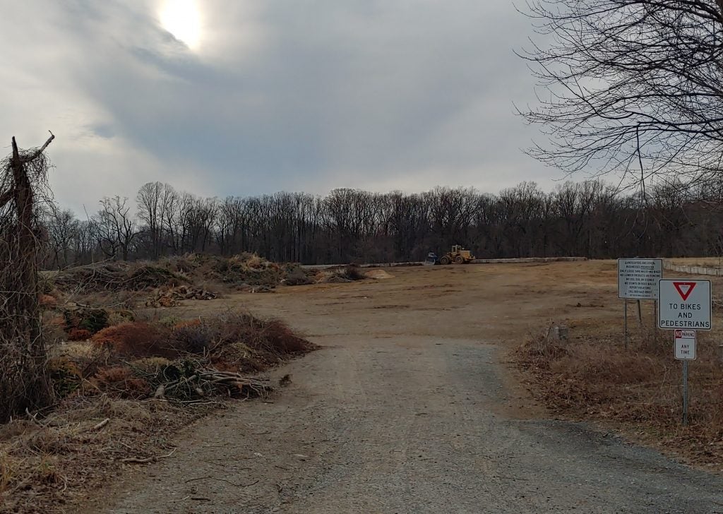 A waste site stretches toward a line of trees under a partly cloudy sky.
