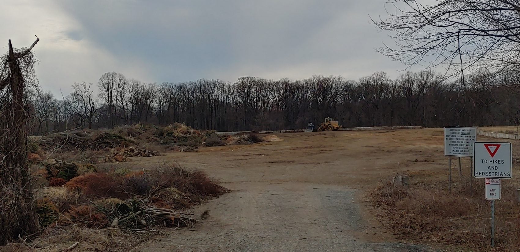 A waste site stretches toward a line of trees under a partly cloudy sky.