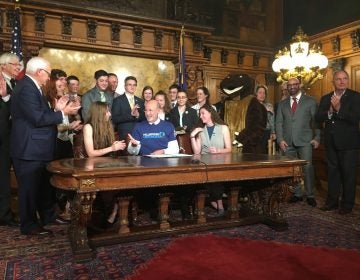 Surrounded by student supporters of the hellbender — and a mascot — Gov. Tom Wolf signs a bill officially designating the salamander as state amphibian. (Katie Meyer/WITF)