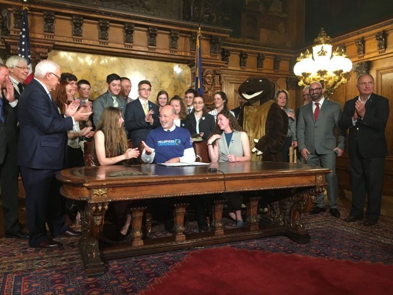 Surrounded by student supporters of the hellbender — and a mascot — Gov. Tom Wolf signs a bill officially designating the salamander as state amphibian. (Katie Meyer/WITF)