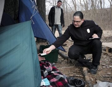 In this Tuesday, Feb. 12, 2019 photo Worcester Police officer Angel Rivera, right, returns a license to an unidentified man as Rivera asks if he has been tested for Hepatitis A at the entrance to a tent where the man spent the night in a wooded area, in Worcester, Mass. (Steven Senne/AP Photo)