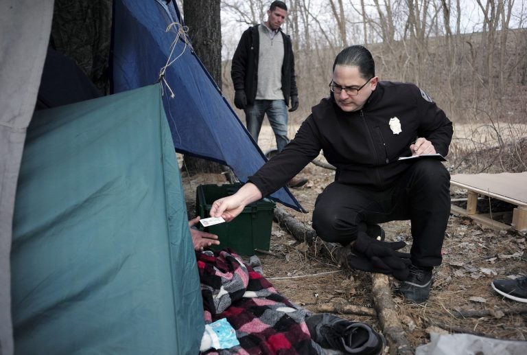 In this Tuesday, Feb. 12, 2019 photo Worcester Police officer Angel Rivera, right, returns a license to an unidentified man as Rivera asks if he has been tested for Hepatitis A at the entrance to a tent where the man spent the night in a wooded area, in Worcester, Mass. (Steven Senne/AP Photo)