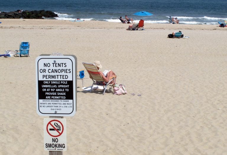 In this Monday, May 20, 2019 photo, beachgoers sit on the sand beyond a sign indicating that smoking is prohibited on the beach in Spring Lake, N.J. A statewide smoking ban is in effect at New Jersey's beaches for the start of the 2019 summer season, although towns can set aside up to 15% as smoking sections. (Wayne Parry/AP Photo)