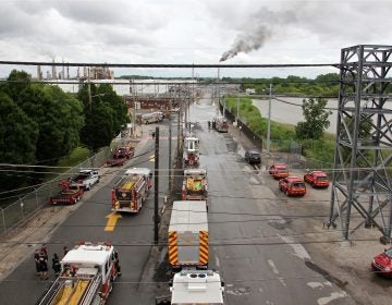 In this file photo, Philadelphia firefighters gather at the entrance to Philadelphia Energy Solutions Refinery, while a large flare burns off fuel to prevent it from feeding the massive fire at the refinery, Friday, June 21, 2019. (Emma Lee/WHYY, file)