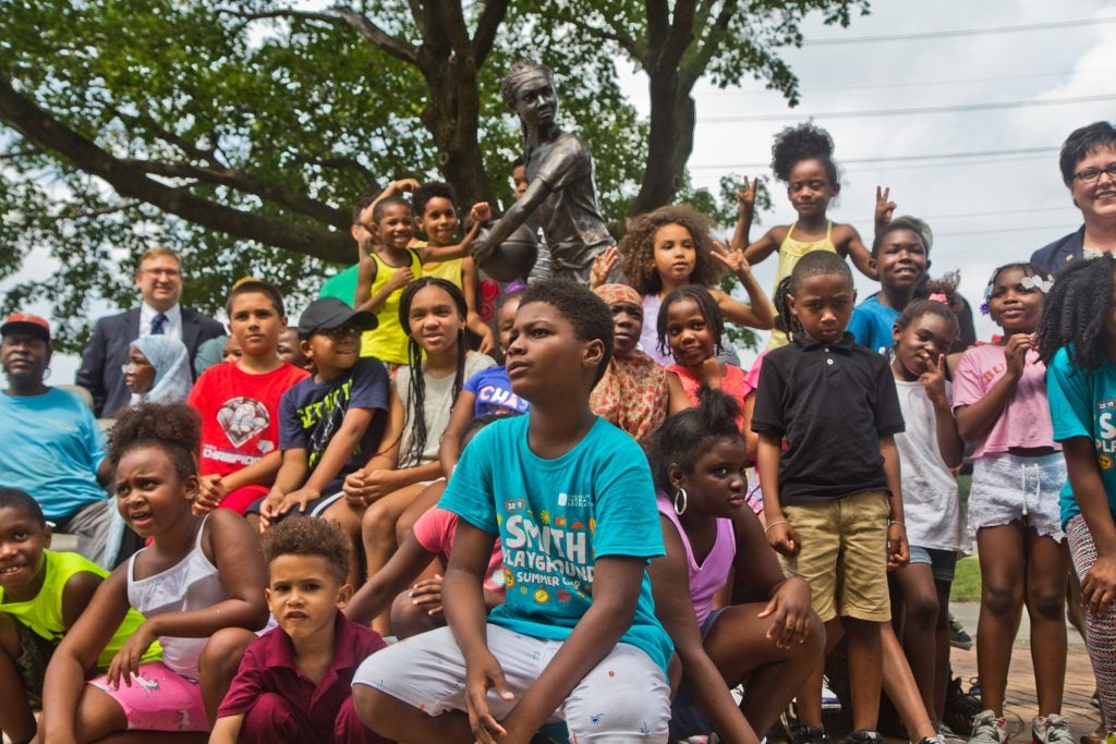 A large group of kids posing for a photo around a statue