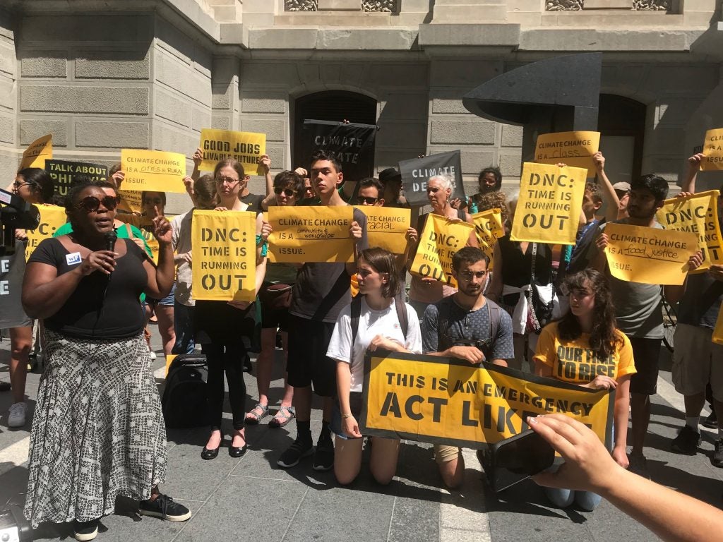 Kendra Brooks speaks outside of City Hall with a microphone in hand. There is a group of people standing next to her and holding signs.