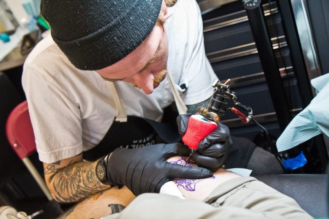 Artist Jacob Des tattoos a cat in a bowtie onto Mike Jenette’s thigh. (Kimberly Paynter/WHYY)