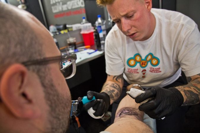 True Hand tattoo artist Chris Fernandez gives a tattoo of the grim reaper holding a bad news sign to Jamie Santoro. (Kimberly Paynter/WHYY)