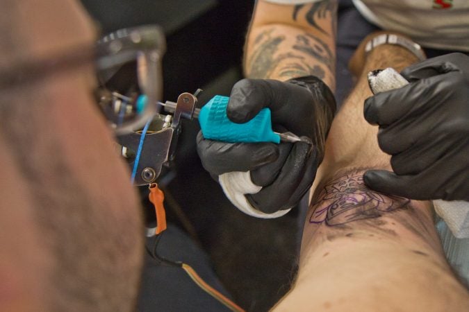 True Hand tattoo artist Chris Fernandez gives a tattoo of the grim reaper holding a bad news sign to Jamie Santoro. (Kimberly Paynter/WHYY)