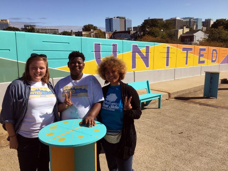 West Side Grows Together's Sarah Lester, volunteer Brenden Cephas and United Neighbors organizer Vanity Constance stand in front of a mural on the Seventh Street bridge over I-95. (Mark Eichmann/WHYY)