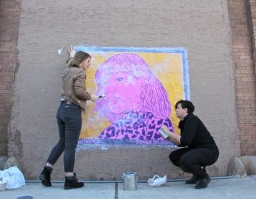 Nicole Nikolich (left) and Symone Salib install Salib's wheat paste portrait of Santigold on a wall of The Filmore. The work is one of 20 in the #SisterlyLove Project, celebrating 20 Philadelphia women during Women's History Month. (Emma Lee/WHYY)