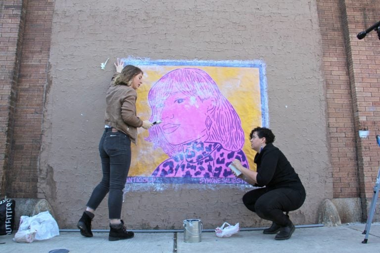 Nicole Nikolich (left) and Symone Salib install Salib's wheat paste portrait of Santigold on a wall of The Filmore. The work is one of 20 in the #SisterlyLove Project, celebrating 20 Philadelphia women during Women's History Month. (Emma Lee/WHYY)