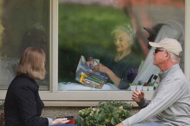 Judie Shape, center, who has tested positive for the new coronavirus, but isn't showing symptoms, opens a care package of art supplies from her daughter and her son-in-law, Tuesday, March 17, 2020, as they talk on the phone and look at each other through a window at the Life Care Center in Kirkland, Wash., near Seattle. (AP Photo/Ted S. Warren)