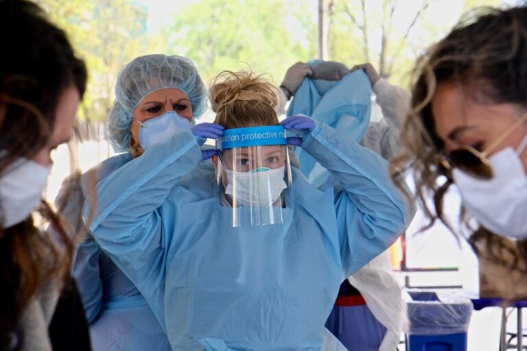 Medical assistant Michelle Gravinese (left) helps her colleague Kayla Clauso put on protective gear before administering COVID-19 tests at a testing site in the Motor Vehicle Commission parking lot in Camden, New Jersey. (Emma Lee/WHYY)