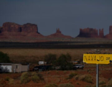In this April 30, 2020, photo, a sign marks Navajo Drive as Sentinel Mesa, homes and other structures in Oljato-Monument Valley, Utah on the Navajo Reservation, stand in the distance. (AP Photo/Carolyn Kaster)