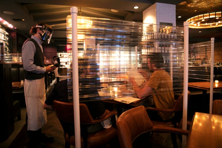 In this June 22, 2020, file photo, wearing a face shield, server Brandon Fricke, left, takes orders from dine-in customers surrounded by protective dividers at Water Grill seafood restaurant in Los Angeles. (AP Photo/Jae C. Hong)