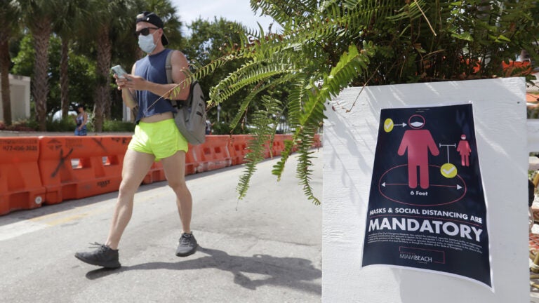 A pedestrian in a mask passes a sign urging people to practice social distancing, on Saturday in Miami Beach, Fla. Just as residents flocked outside to enjoy the Fourth of July, states such as Florida were reporting skyrocketing numbers of confirmed coronavirus cases. (Wilfredo Lee/AP Photo)
